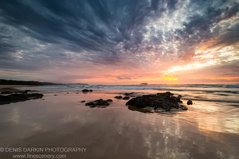 Streaked dawn clouds above a wet mirror beach, dark jagged rocks in the foreground, and a distant headland on the Queensland coast.