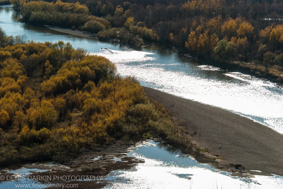 Sunlit gravel bar on the Iman River in late autumn, with golden forest banks and shallow pools reflecting the sky.