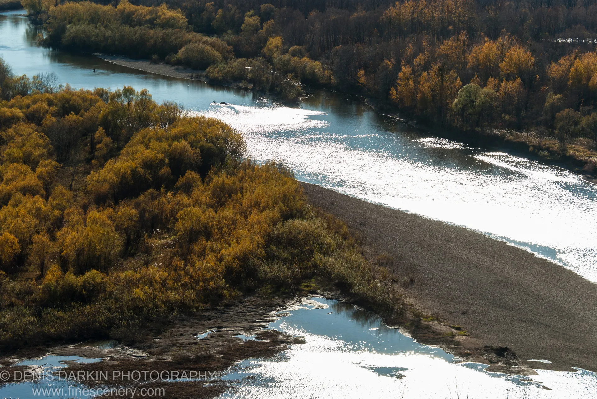 Sunlit gravel bar on the Iman River in late autumn, with golden forest banks and shallow pools reflecting the sky.