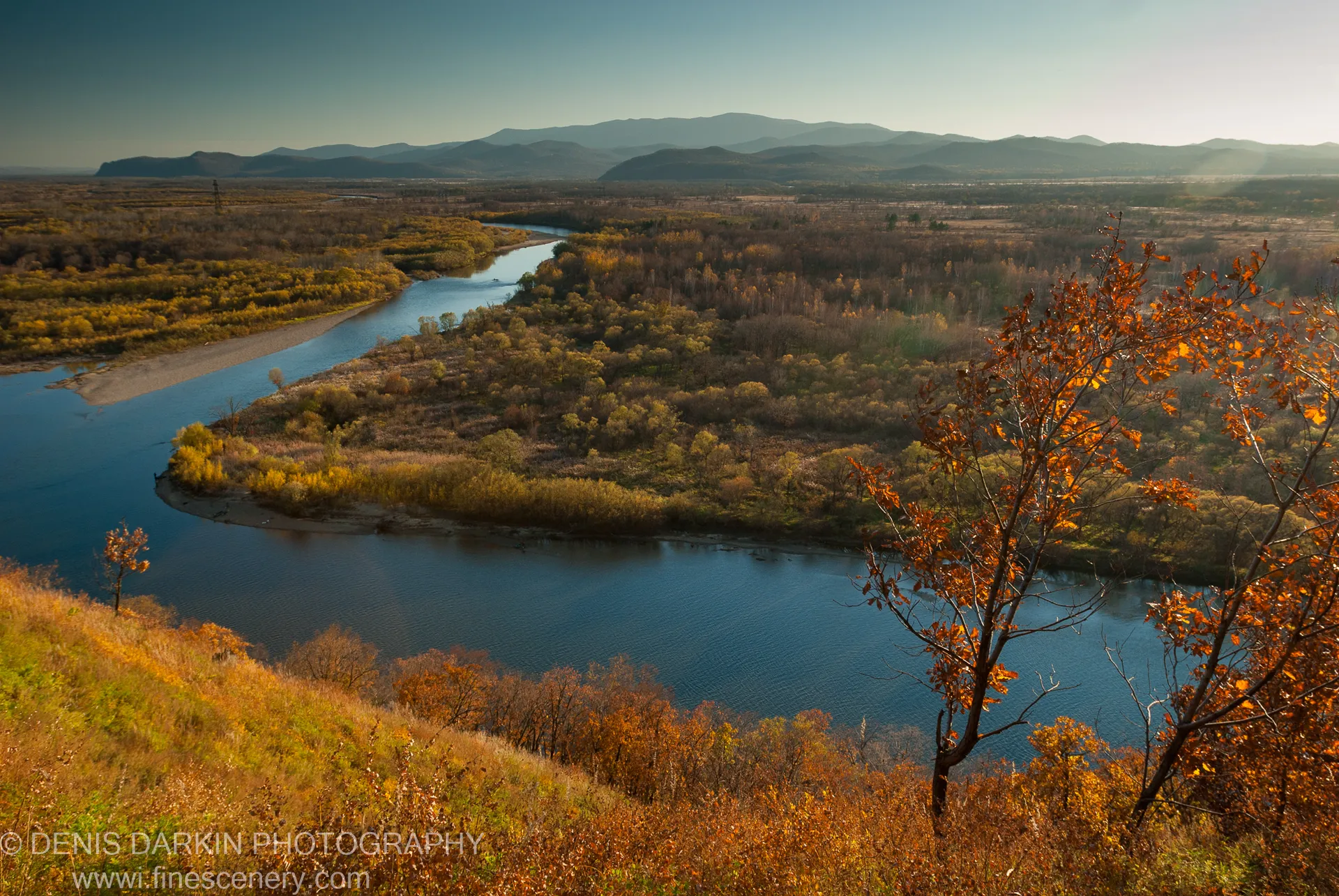 Textured autumn slope and scattered trees in the foreground, with the Iman River and layered hazy mountains receding into the distance.