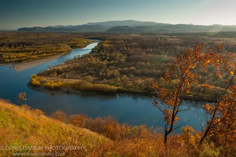 Textured autumn slope and scattered trees in the foreground, with the Iman River and layered hazy mountains receding into the distance.