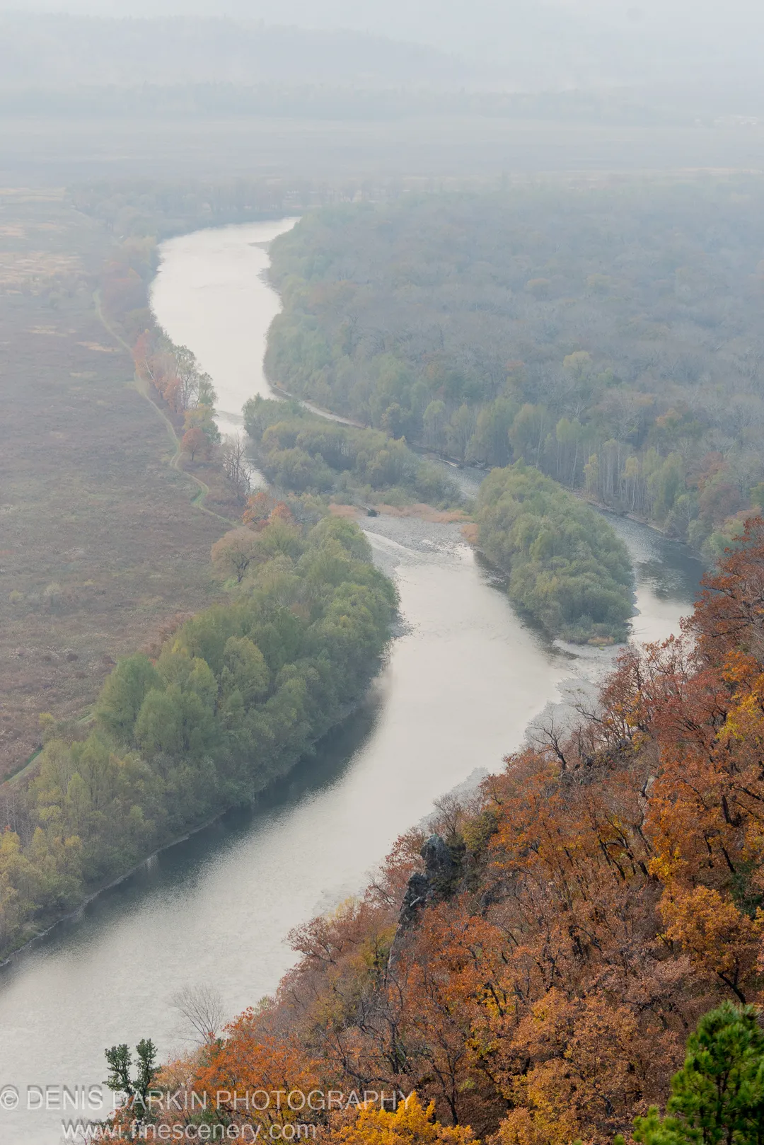 High-angle view of the Iman River winding through an autumn valley, with vibrant foliage in the foreground and forested banks fading into haze.