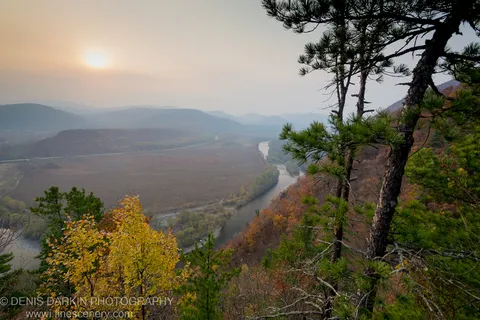 Yellow autumn foliage and a pine silhouette border a valley view of the Iman River winding below hazy mountain ranges.