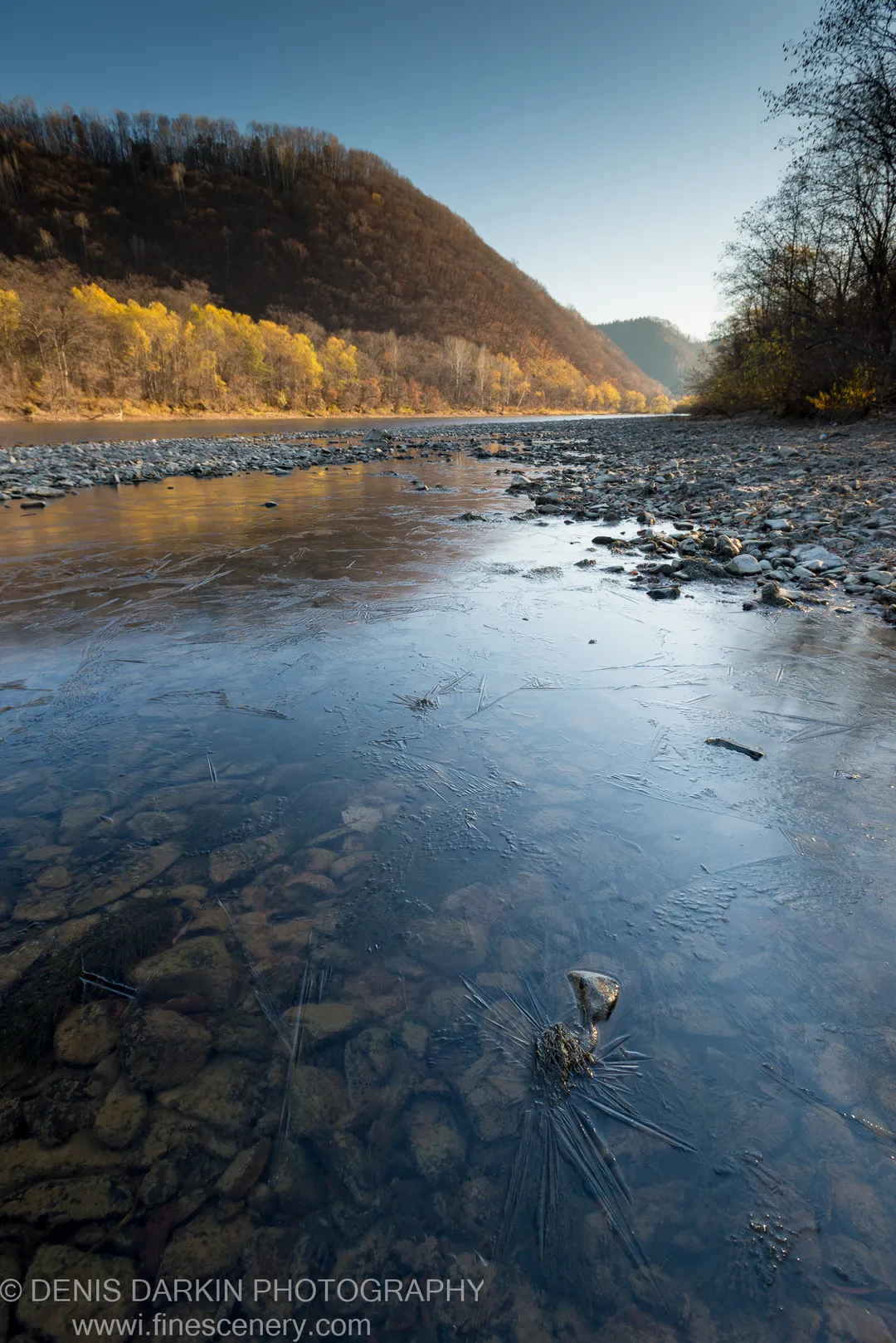 Ice fractured across the shallows of the Iman River, revealing smooth stones beneath, with an autumn hillside rising beyond.