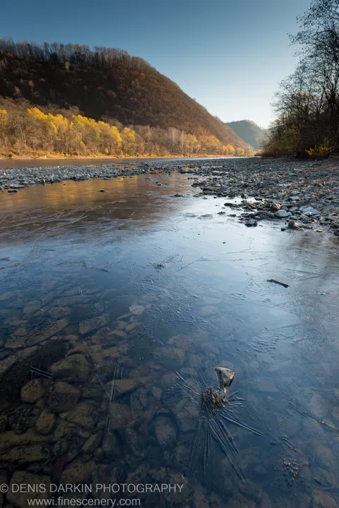 Ice fractured across the shallows of the Iman River, revealing smooth stones beneath, with an autumn hillside rising beyond.