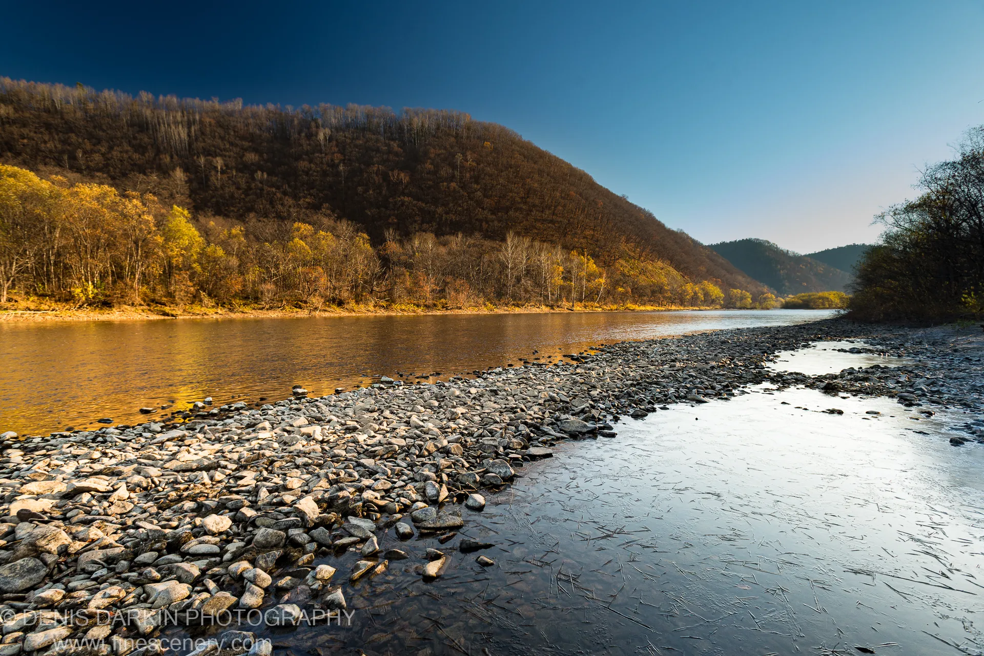 A wide gravel bar on the Iman River catches morning light, the far hillside showing late-autumn foliage against a deep blue sky.