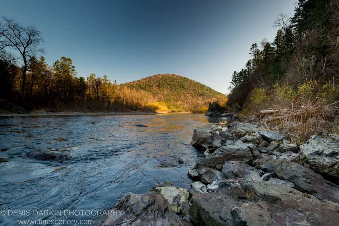 Wide river valley in late afternoon light, rocky shoreline in the foreground and golden-lit forest hillside across the water, Iman River, October 2013.