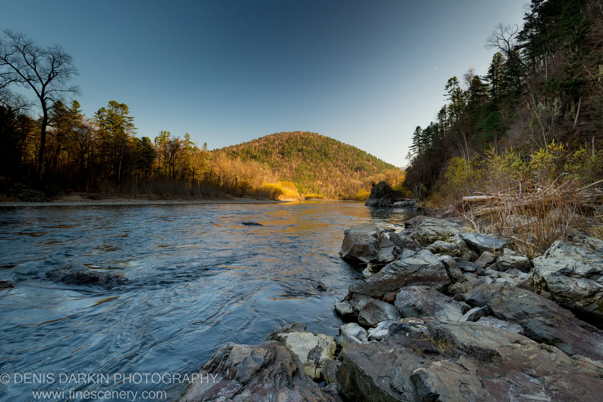 Wide river valley in late afternoon light, rocky shoreline in the foreground and golden-lit forest hillside across the water, Iman River, October 2013.