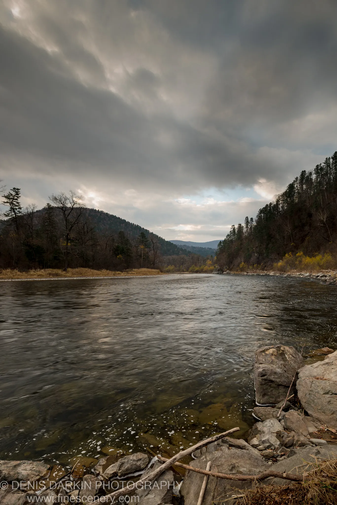 Large boulders and a fallen branch anchor the foreground at the Iman River, storm clouds above bare deciduous trees and dark mixed forest ridges.