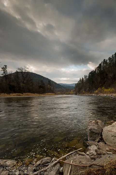 Large boulders and a fallen branch anchor the foreground at the Iman River, storm clouds above bare deciduous trees and dark mixed forest ridges.