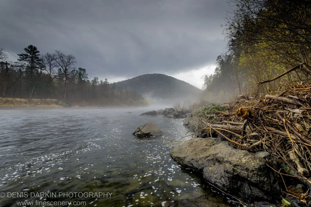Fallen timber on a rocky bank of the Iman River, mist rising from the water beneath an overcast sky.