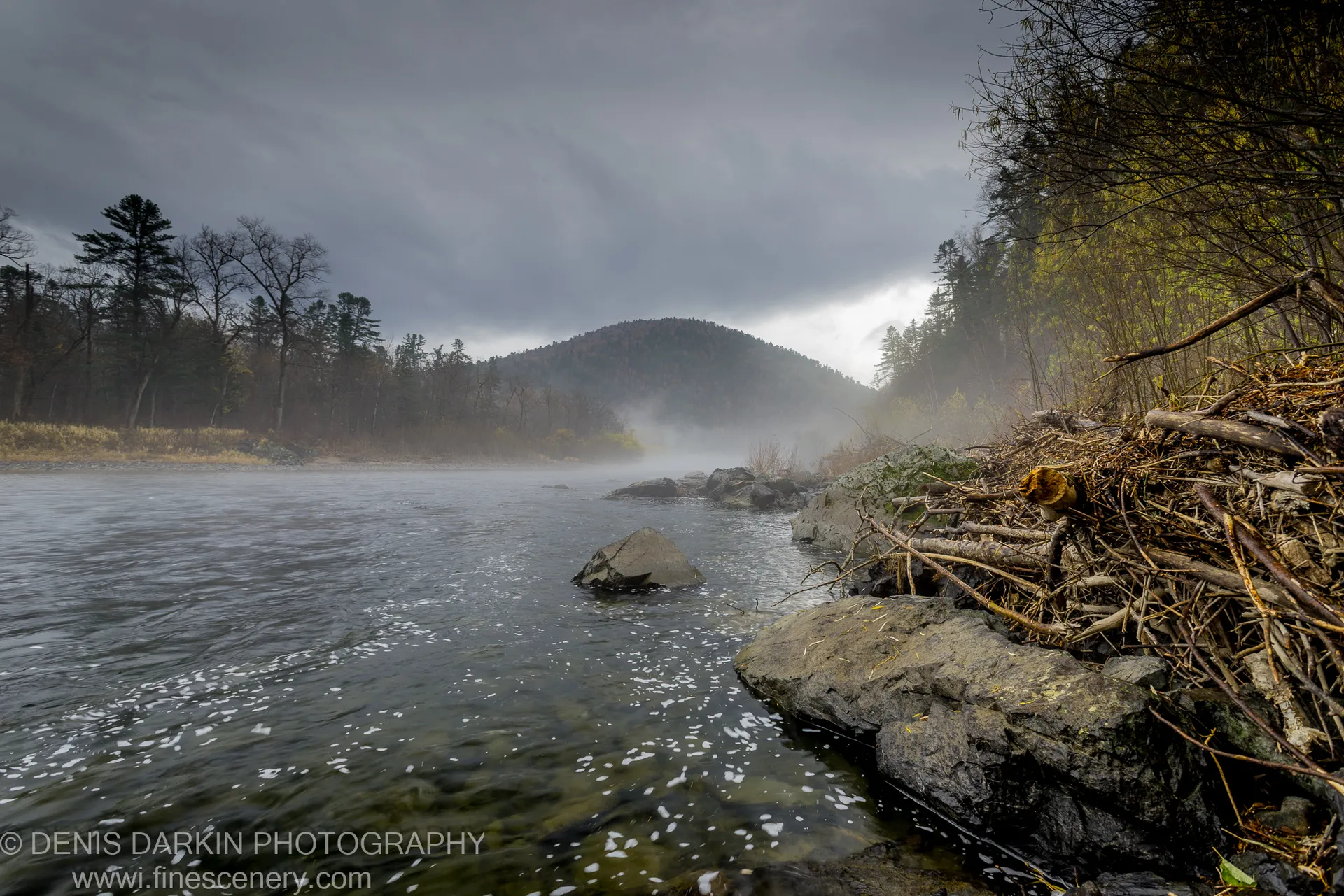 Fallen timber on a rocky bank of the Iman River, mist rising from the water beneath an overcast sky.