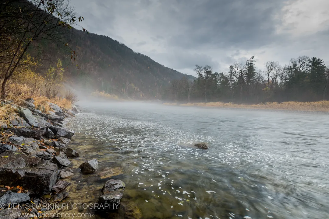 Jagged dark rock shoreline in the foreground with submerged stones visible through shallow water, mist rising from a steep forested hillside across the river.