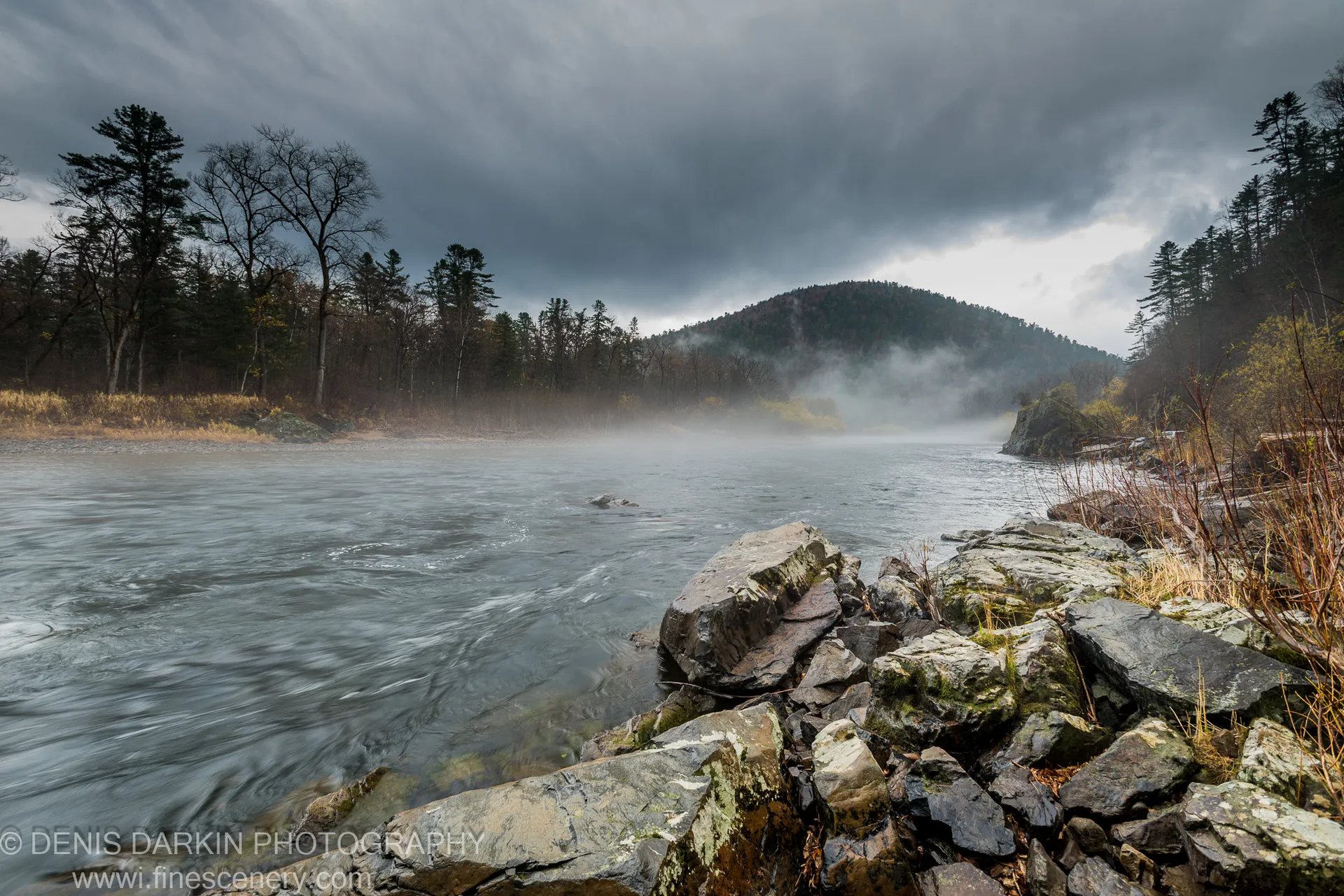 Moss-stained boulders line the near bank of a wide river, mist hanging low over the water, heavy storm clouds above mixed deciduous and conifer ridges.