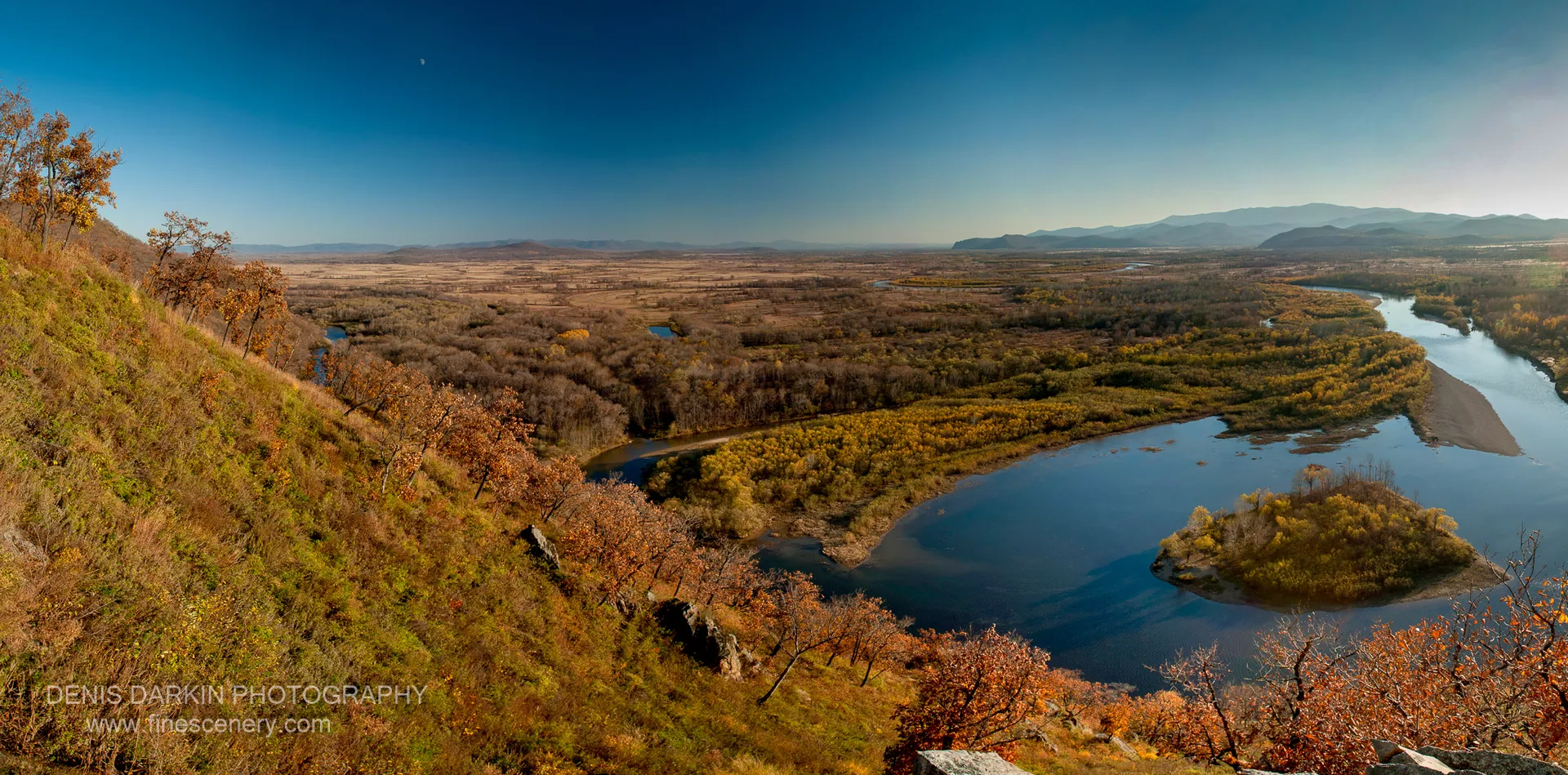 Steep grassy slope with autumn trees above the Iman River valley, a faint crescent moon in the blue sky above.