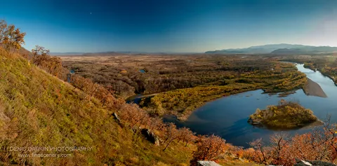 Steep grassy slope with autumn trees above the Iman River valley, a faint crescent moon in the blue sky above.