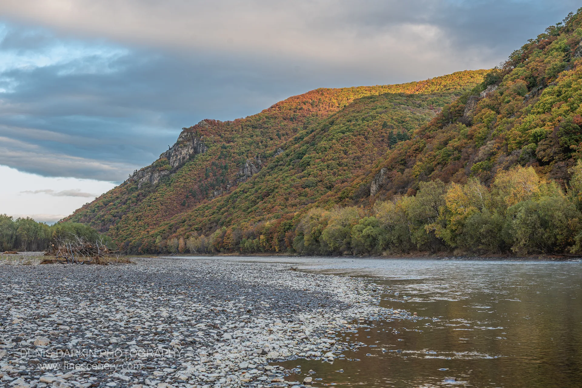 Wide river with driftwood shore in foreground and a forested ridge lit at the crest, Primorsky Krai, October 2019.