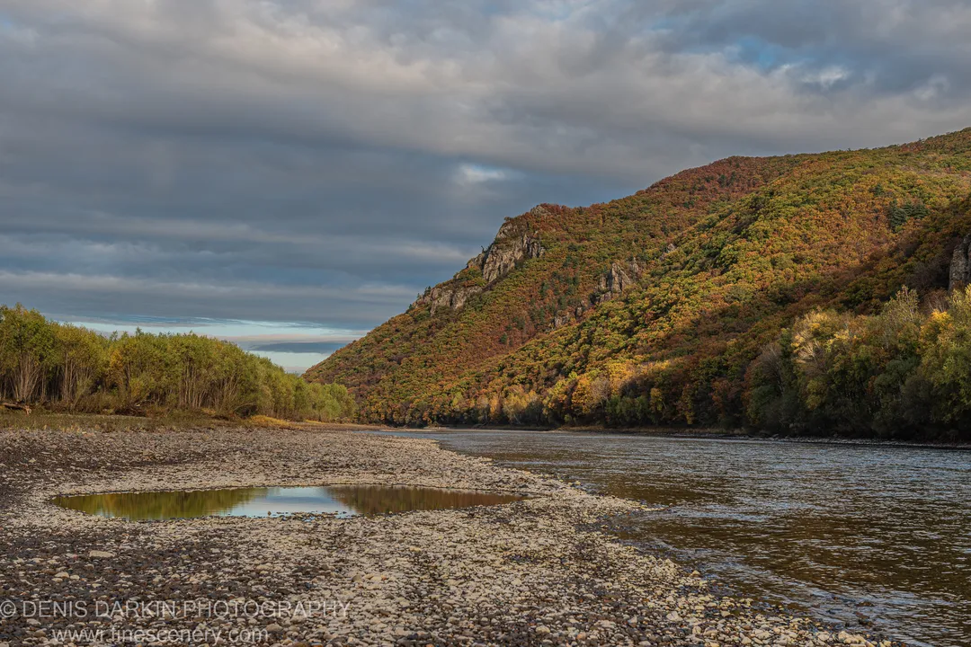Still pool on a gravel bank reflecting a steep foliage hillside, warm autumn light, Primorsky Krai, October 2019.