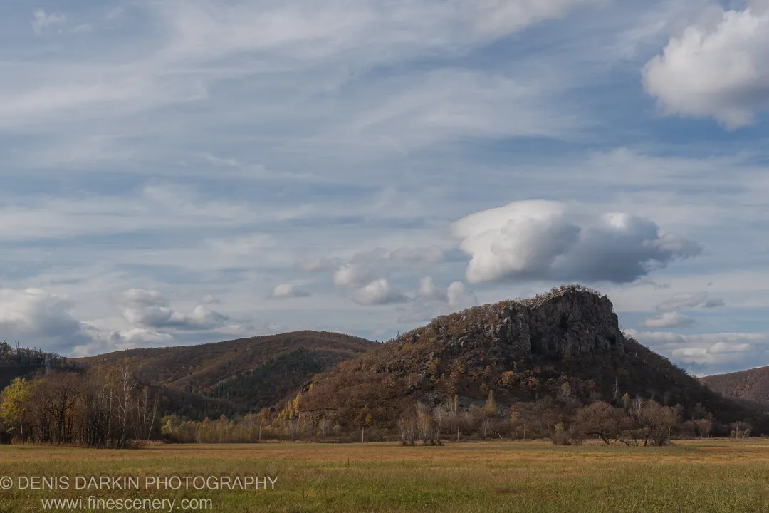 A craggy rocky hill rising from a flat autumn meadow in Primorsky Krai, cirrus clouds above the summit.