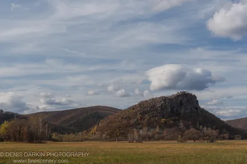 A craggy rocky hill rising from a flat autumn meadow in Primorsky Krai, cirrus clouds above the summit.
