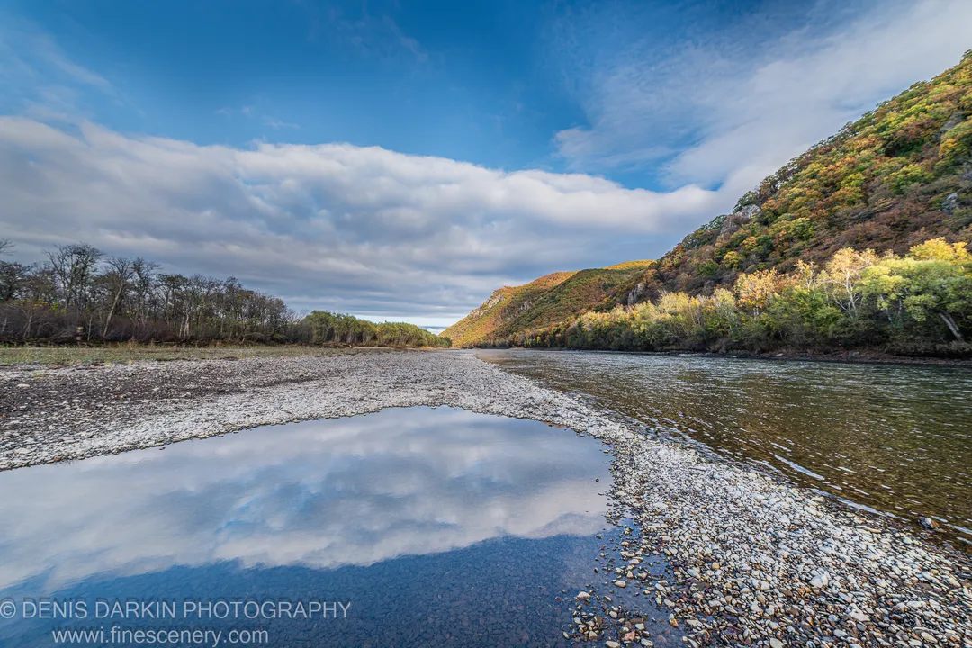 Still pool on a gravel bar mirroring blue sky and wispy clouds, autumn-colored forest on the steep far bank, Primorsky Krai, October 2019.