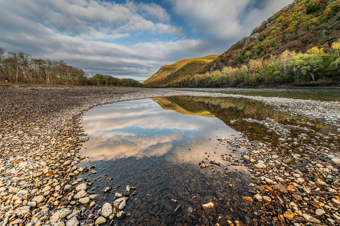 A shallow pool in a rocky riverbed reflects clouds and autumn-colored hillsides in the Russian Far East on an October morning.