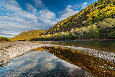 Wide river below a hillside in vibrant autumn color, shallow pool reflecting clouds and trees over smooth submerged stones, Primorsky Krai, October 2019.