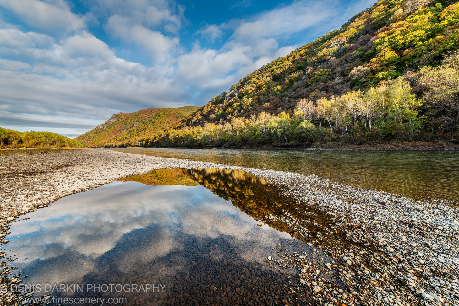 River flowing past a sunlit gravel bar in Primorsky Krai, autumn hillside in yellow and green reflected in a still pool.