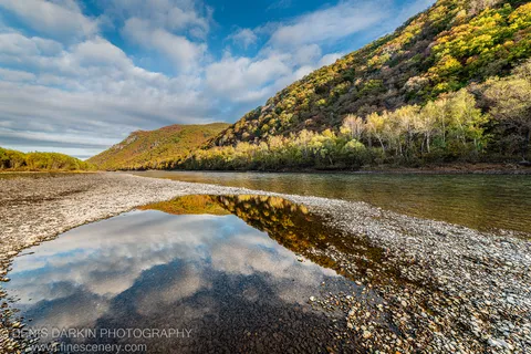 River flowing past a sunlit gravel bar in Primorsky Krai, autumn hillside in yellow and green reflected in a still pool.