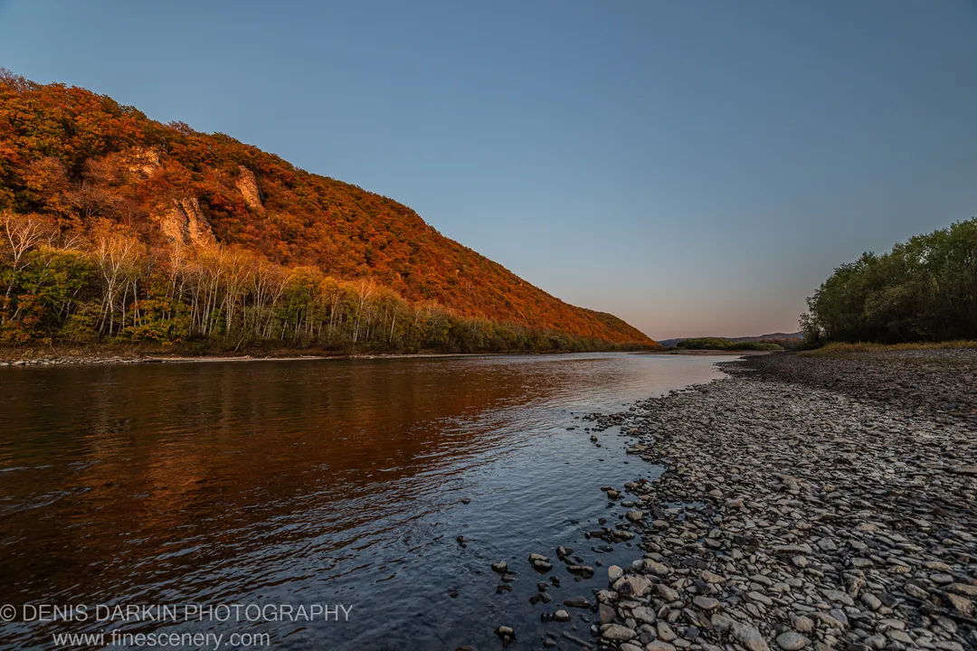 Rocky pebble shore on a wide river at sunset, Primorsky Krai. Autumn hillside in warm orange and yellow on the far bank.