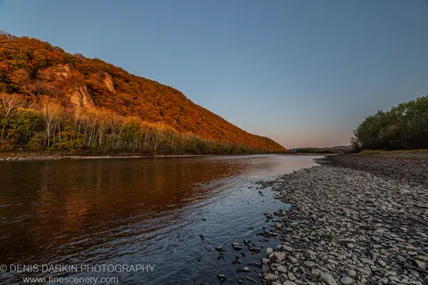 Rocky pebble shore on a wide river at sunset, Primorsky Krai. Autumn hillside in warm orange and yellow on the far bank.