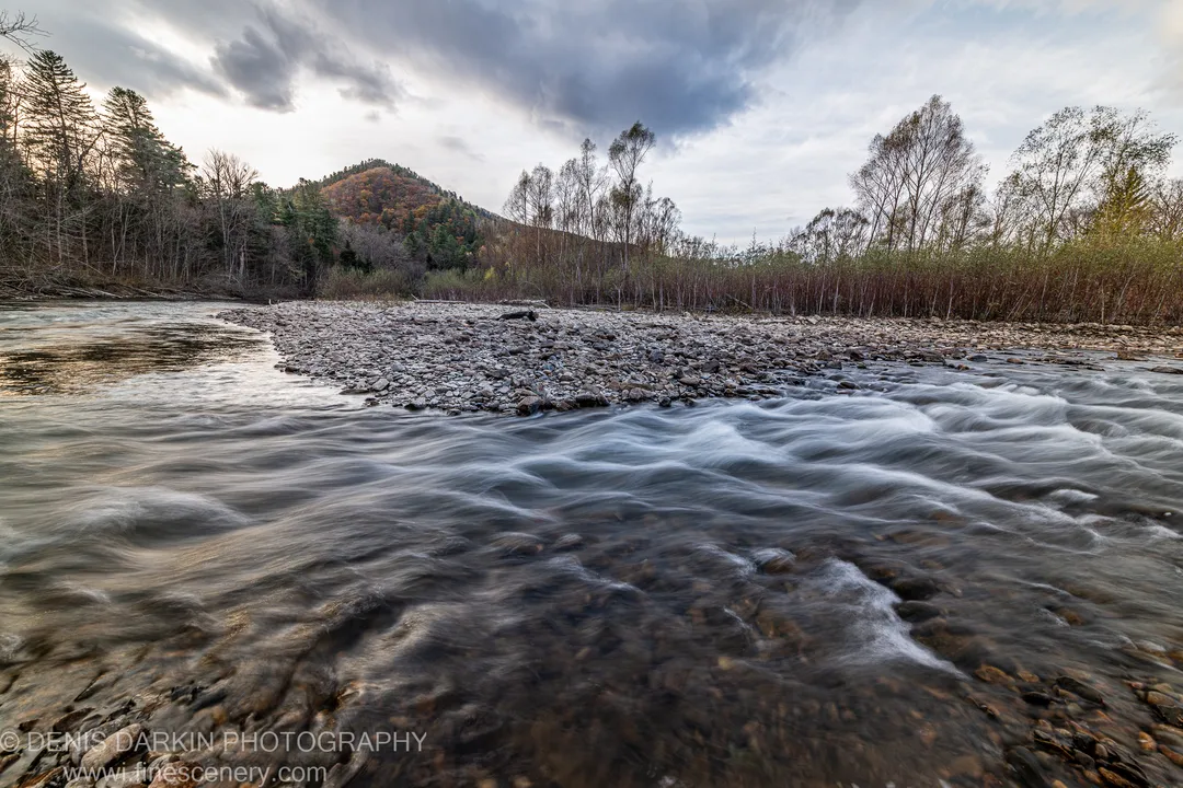 Long-exposure river view in Primorsky Krai with water flowing over submerged stones toward a wide gravel bar beneath dark autumn clouds.