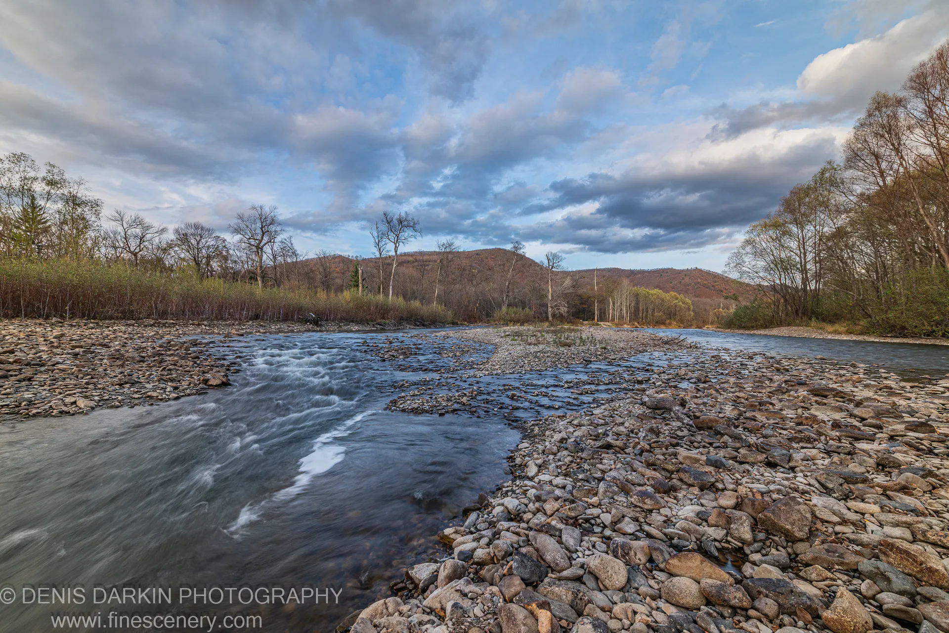 Scattered rounded stones in the foreground with smooth flowing water and rolling autumn hills in the background under a cloudy sky, Primorsky Krai.