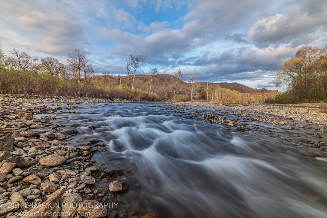 Rounded river pebbles in the foreground, bare autumn trees behind, and a distant wooded ridge under blue sky with scattered clouds, Primorsky Krai.