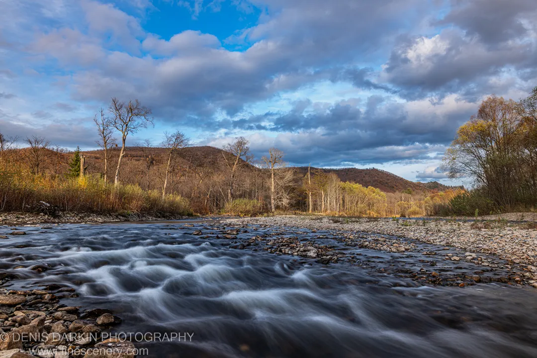 Long-exposure water rushes over smooth river stones at a gravel bar, with rolling hills in autumn color in the background, Russian Far East.