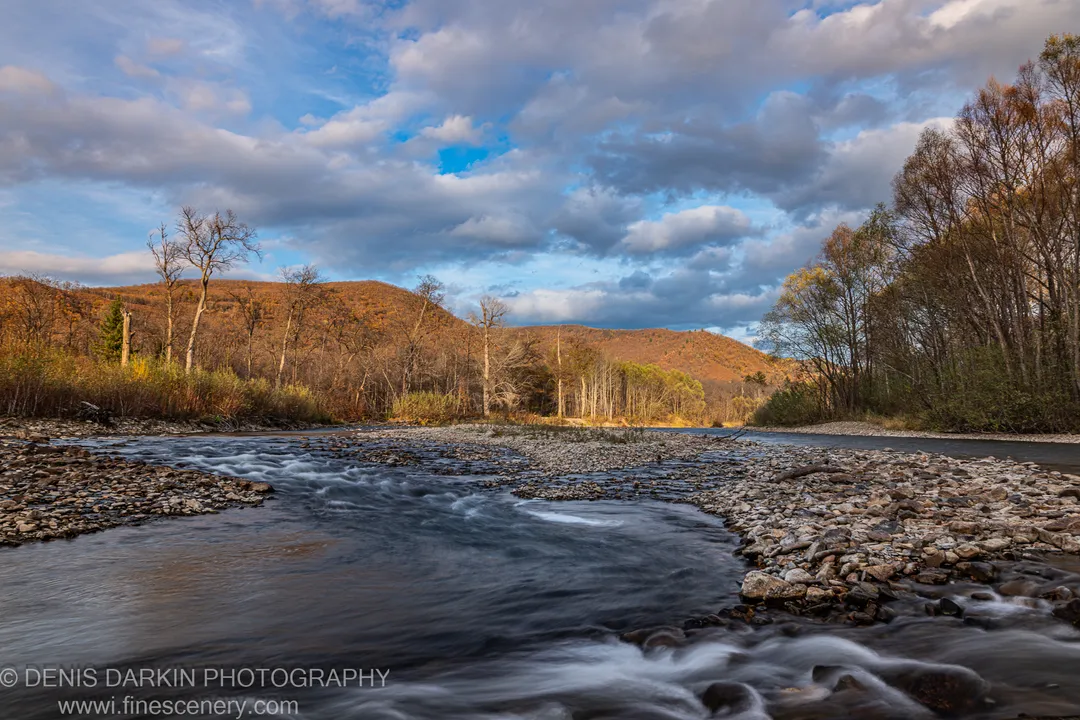 Wide river over a rocky bed with long-exposure water forming white ribbons, autumn hills, and a cloud-marked sky, Primorsky Krai.