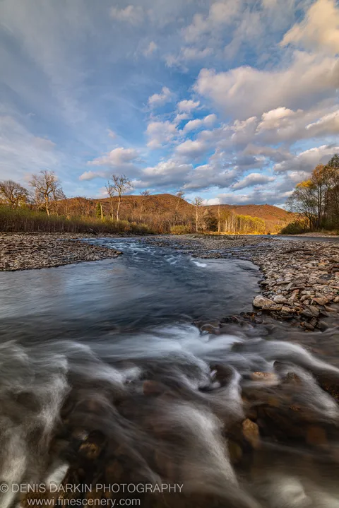 Water moves over a cobbled riverbed at golden hour, with a deciduous hillside in full autumn color rising in the background, Russian Far East.
