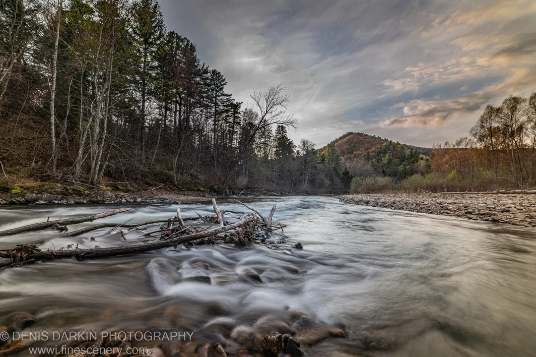River flowing past fallen logs in the foreground, with streaked clouds and a distant autumn hillside beyond, Primorsky Krai.