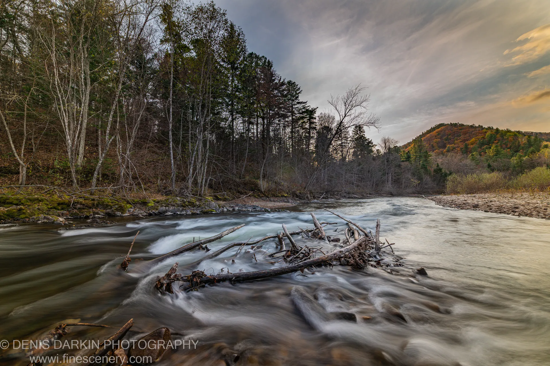 Long-exposure forest stream over submerged timber, with mossy banks, bare deciduous trees, and a distant hillside in autumn foliage, Primorsky Krai.