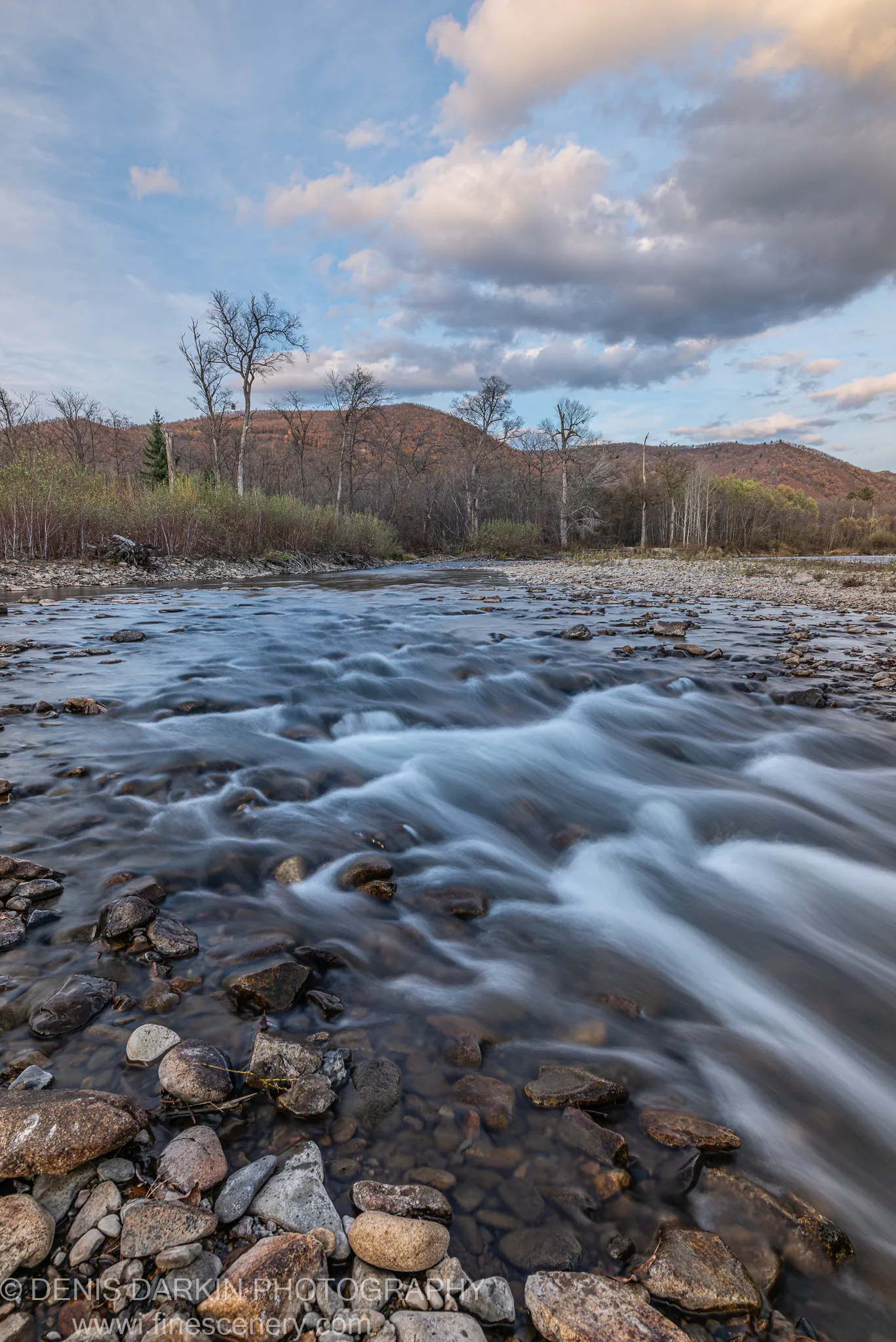 Smooth water over dark river stones leading toward distant hills in late autumn foliage under textured clouds, wide-angle view, Primorsky Krai.