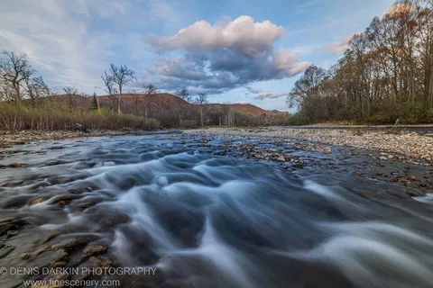 White water flows over dark stones on a wide Primorsky Krai river, backed by reddish autumn hills and textured clouds above tree-lined banks.