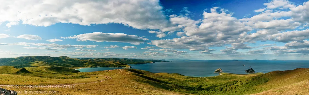 Rolling grass-covered hills and a winding dirt track descend toward a sheltered bay with turquoise water, Astafyeva Bay, Primorsky Krai.