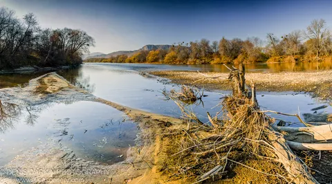 Tangled roots and driftwood sit on muddy riverbanks in the foreground, the Suchan River reflecting autumn-covered hills in the background.