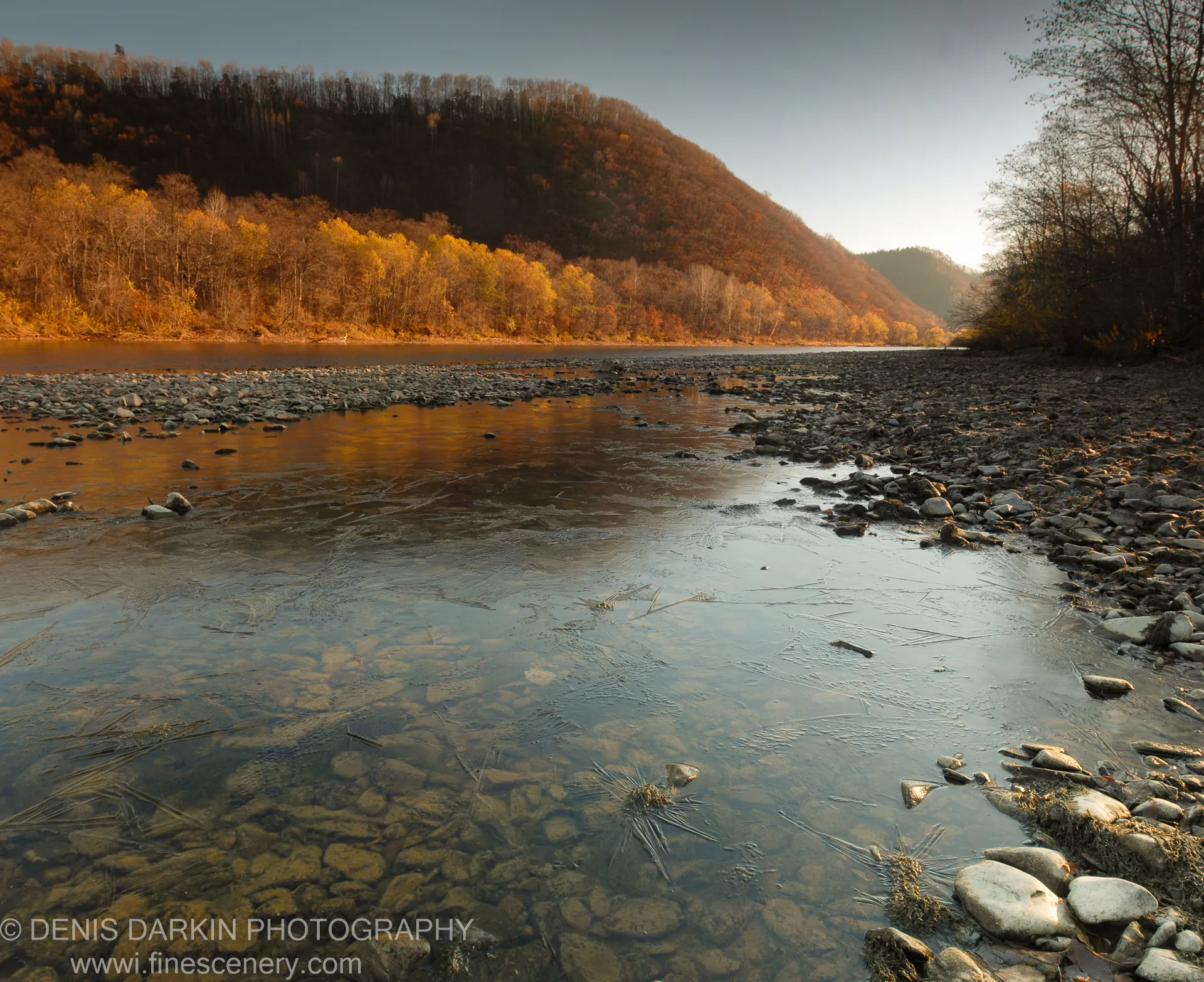 Dark pebbles line a jagged shoreline in the foreground, calm blue water extending toward a rugged mountain peak in the background at golden hour.