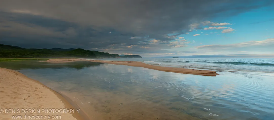 A long sandbar separates a calm reflective inlet from the open ocean at Tachingouza Bay, with storm clouds above forested hills and blue sky beyond.
