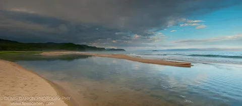A long sandbar separates a calm reflective inlet from the open ocean at Tachingouza Bay, with storm clouds above forested hills and blue sky beyond.