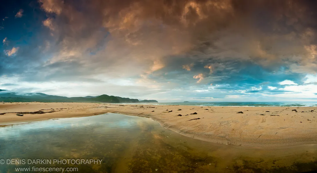 Warm sunset clouds over dark coastal hills and the ocean at Tachingouza Bay, with wet sand and shallow water in the foreground, July 2010.