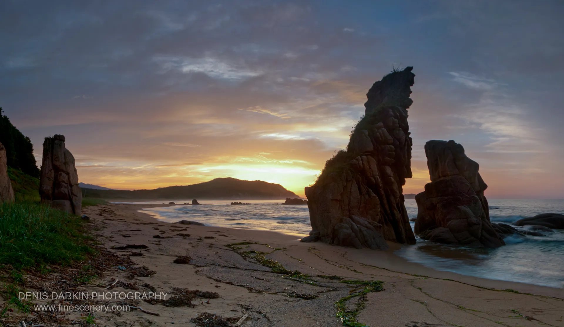 Large weathered sea stacks on a seaweed-scattered shore at Tachingouza Bay, with a warm morning sun low on the horizon.