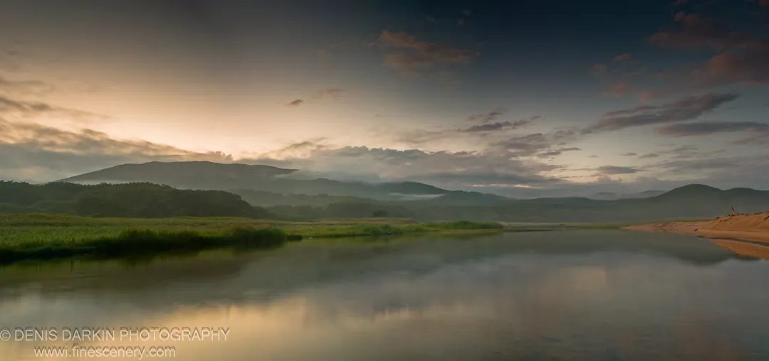 Calm delta waters at Tachingouza Bay reflect a sky gradient from amber to slate blue, with green marshland on the left bank and hazy mountains beyond.