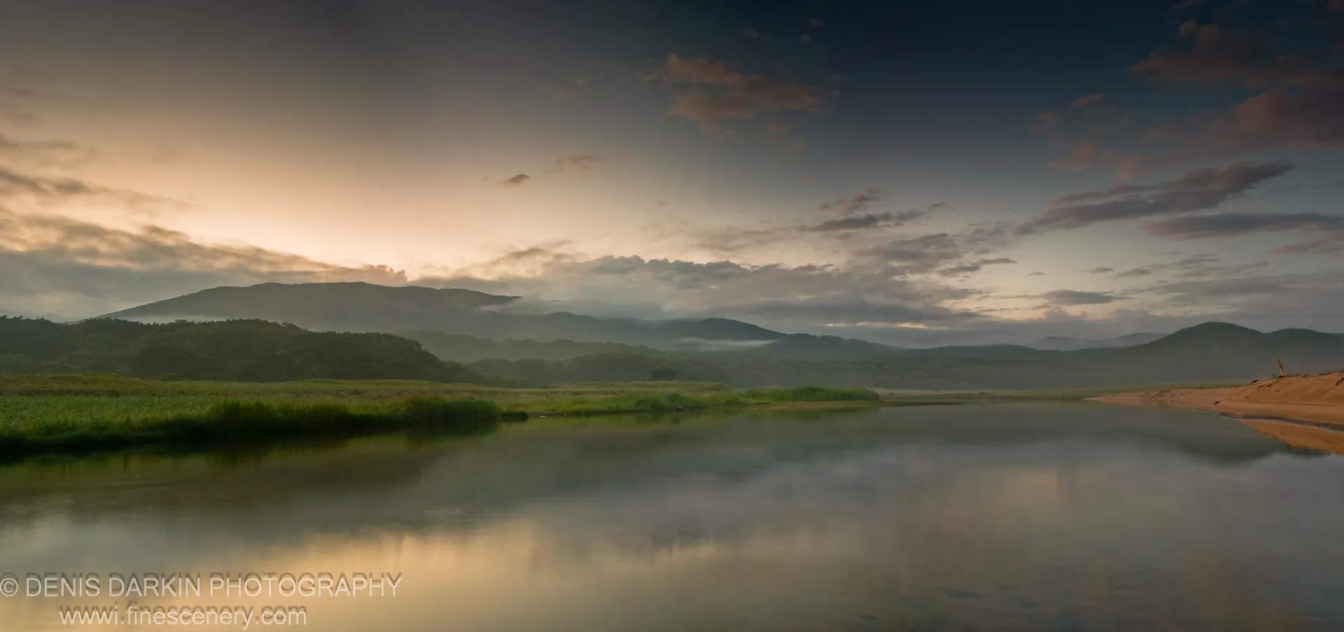 Calm delta waters at Tachingouza Bay reflect a sky gradient from amber to slate blue, with green marshland on the left bank and hazy mountains beyond.